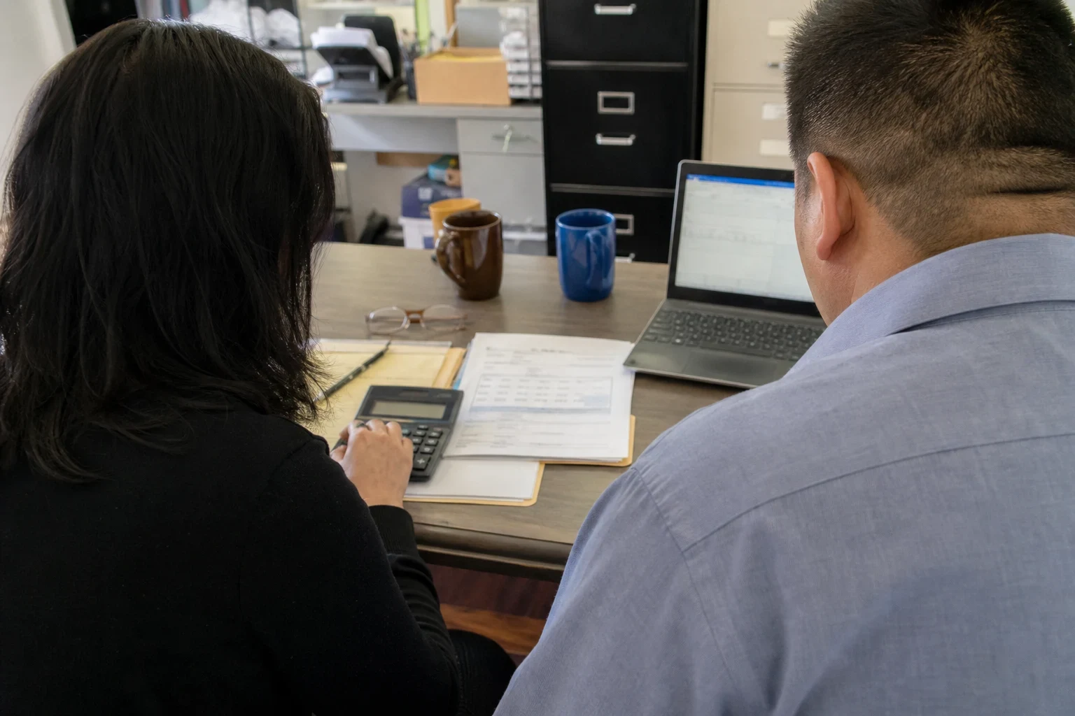 Two people seen from behind at an office desk reviewing printed statements, one using a calculator beside a notepad, glasses, mugs and an open laptop near filing cabinets