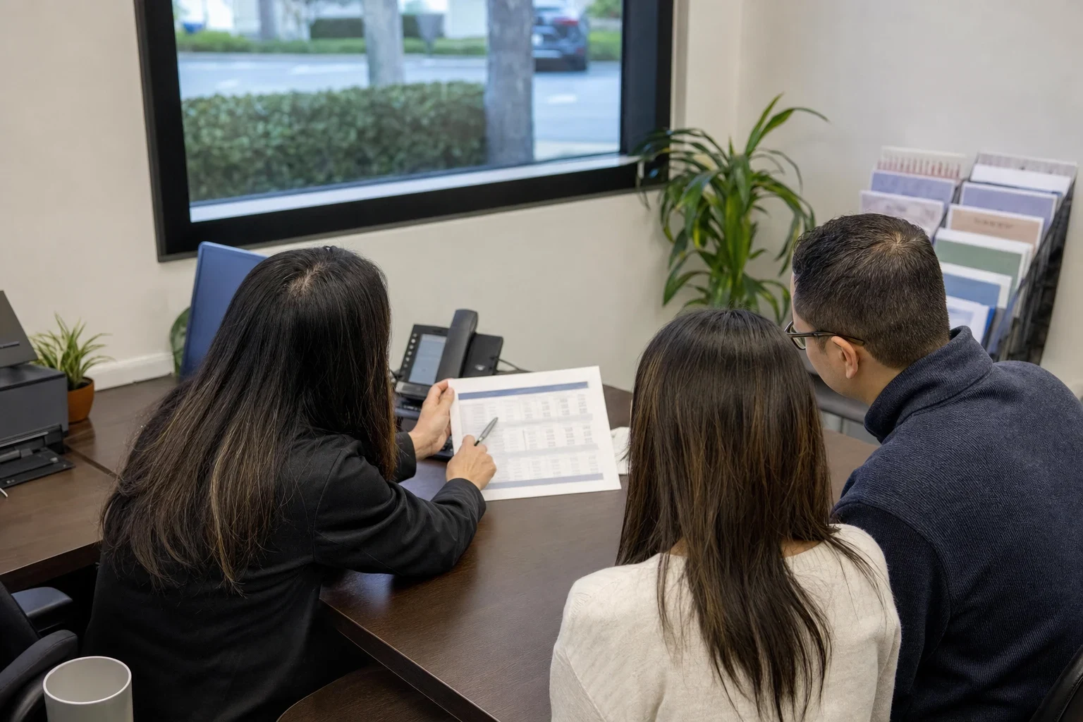 Insurance agent at an office desk pointing a pen at a printed comparison sheet for a couple seated beside her, with a window, plant and brochure rack behind